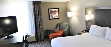 A hotel room with a large bed, a desk with a television, a chair, a small table, and a lamp.