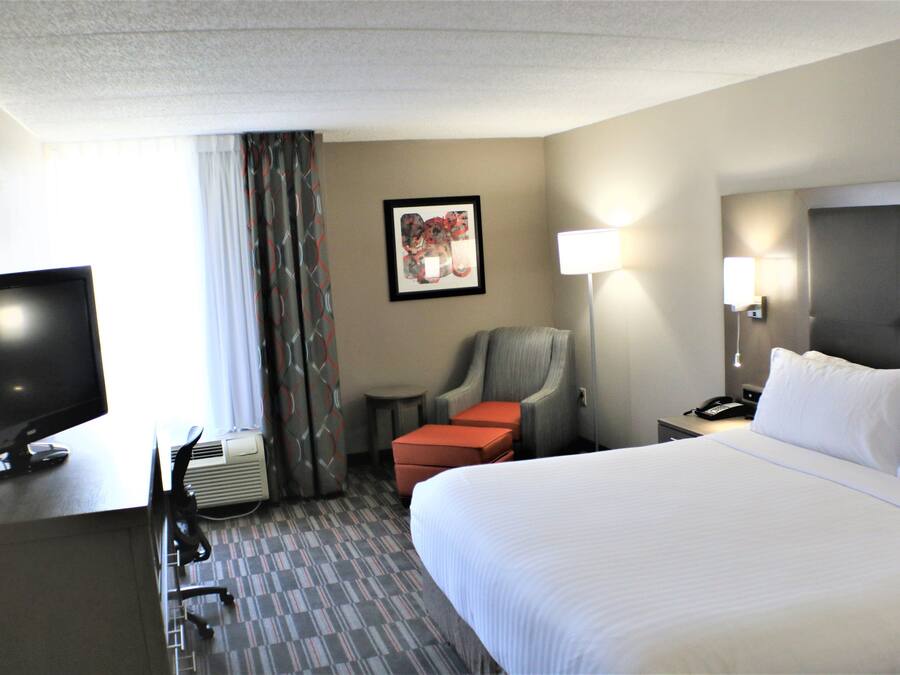 A hotel room with a large bed, a desk with a television, a chair, a small table, and a lamp.