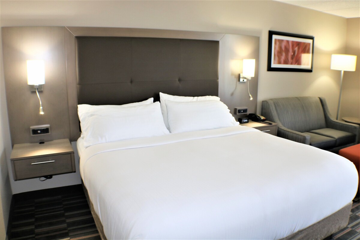 A hotel room with a large bed, a nightstand, a chair, and a wall-mounted lamp.