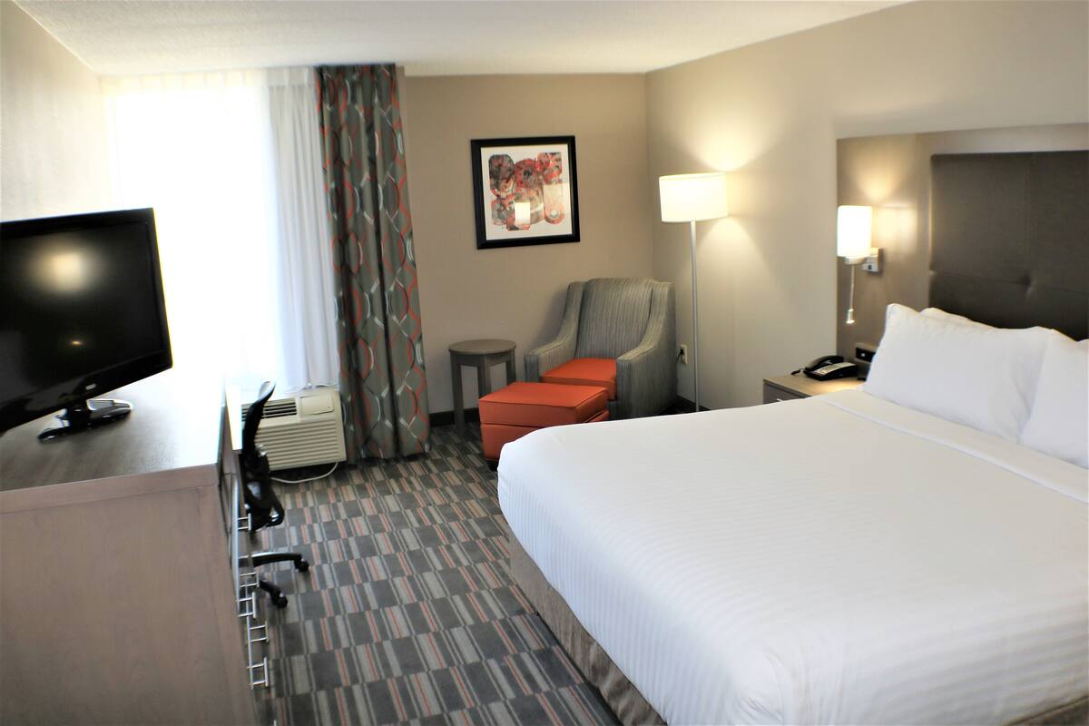 Ein Hotelzimmer mit einem Bett, einem Schreibtisch mit Computer, einem Sessel, einer Lampe und einem Fernseher.