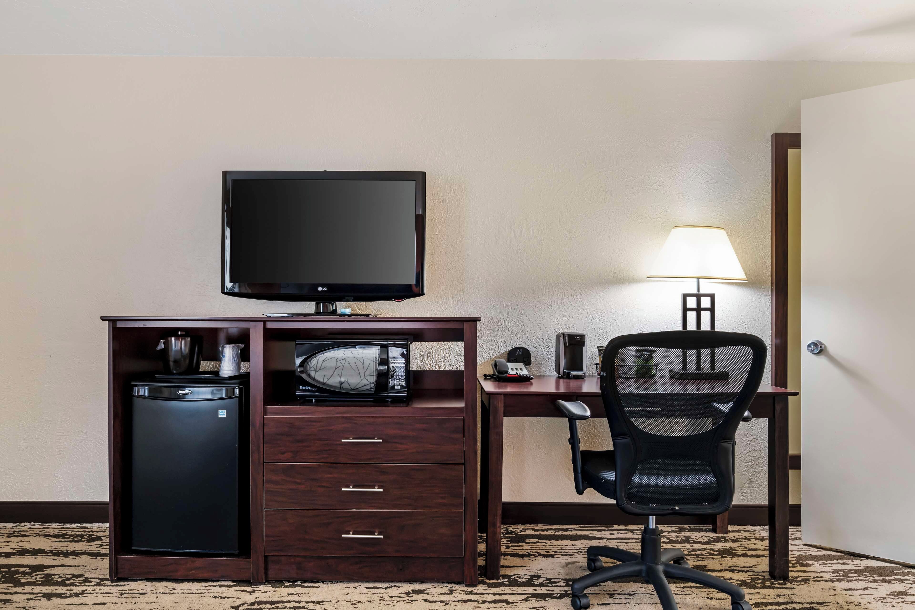 A hotel room with a desk, a television, a microwave, a coffee maker, a chair, and a lamp.