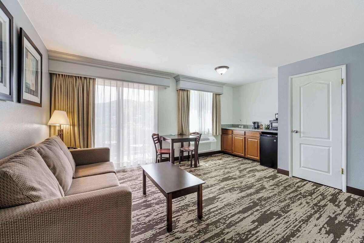 A hotel room with a sofa, a dining table with chairs, a kitchen area with a dishwasher, and a door.
