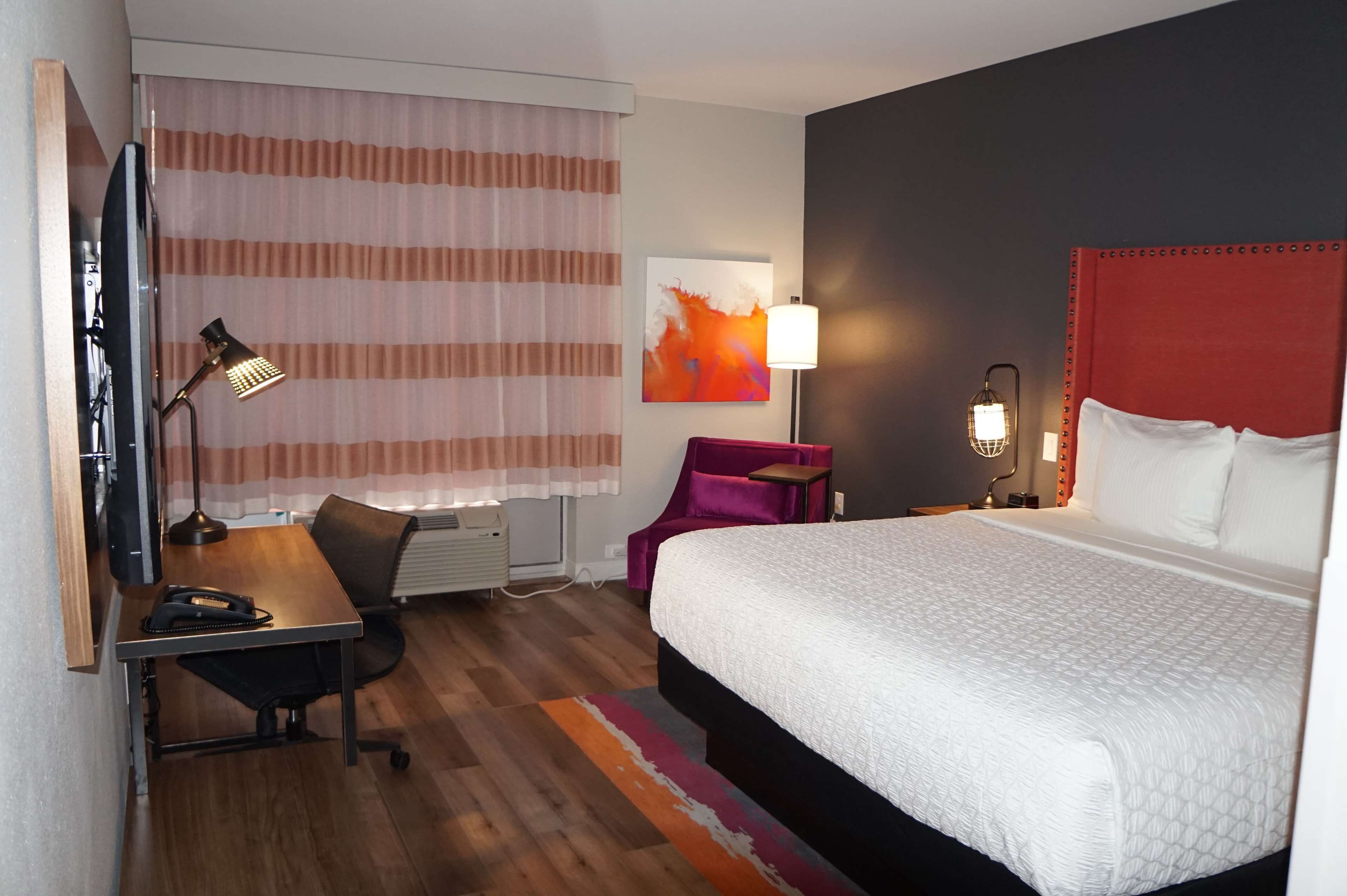 A hotel room with a bed, desk, chair, lamp, and artwork on the wall.