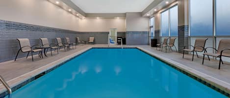 A swimming pool with chairs and a glass door, surrounded by large windows and a tiled wall.