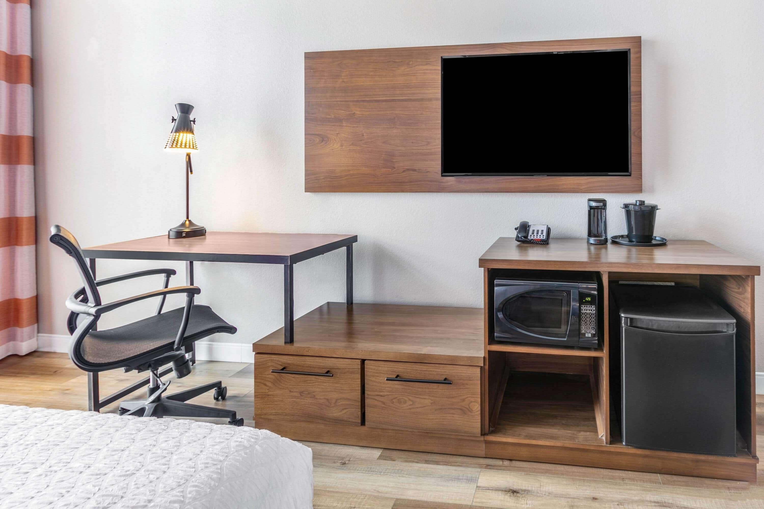 Habitación de hotel compacta con cama, escritorio y silla, televisor montado en la pared, cafetera, microondas y refrigerador.