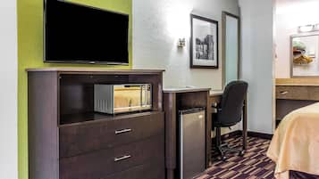 A hotel room with a dark wooden desk, a microwave, a refrigerator, a black office chair, a bed with a beige cover, a framed picture on the wall, and a television mounted above the desk.