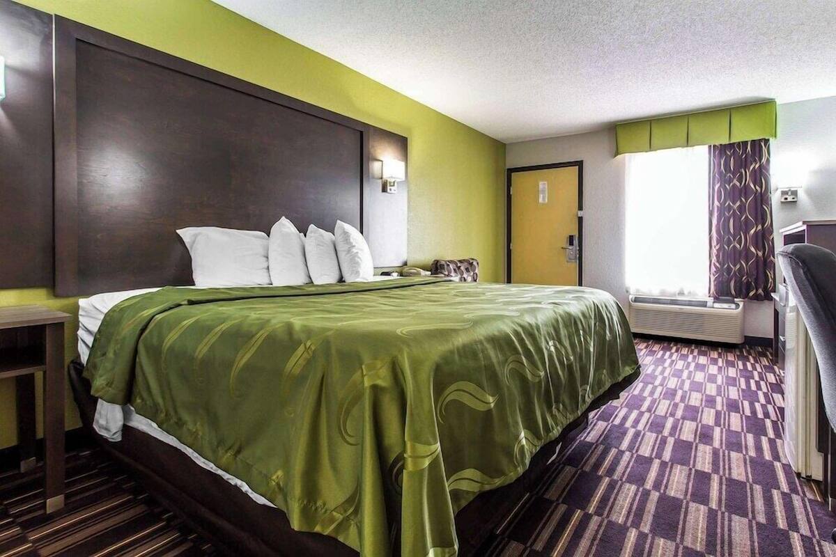 Una habitación de hotel con una cama grande, una mesita de noche, una silla, una ventana con cortinas y una puerta.