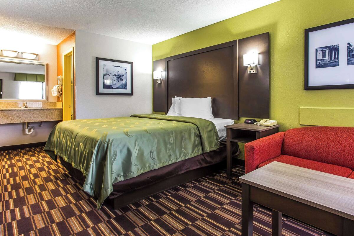Habitación de hotel con cama, sillón rojo, mesita, mesita de noche, espejo y un cuadro en la pared.