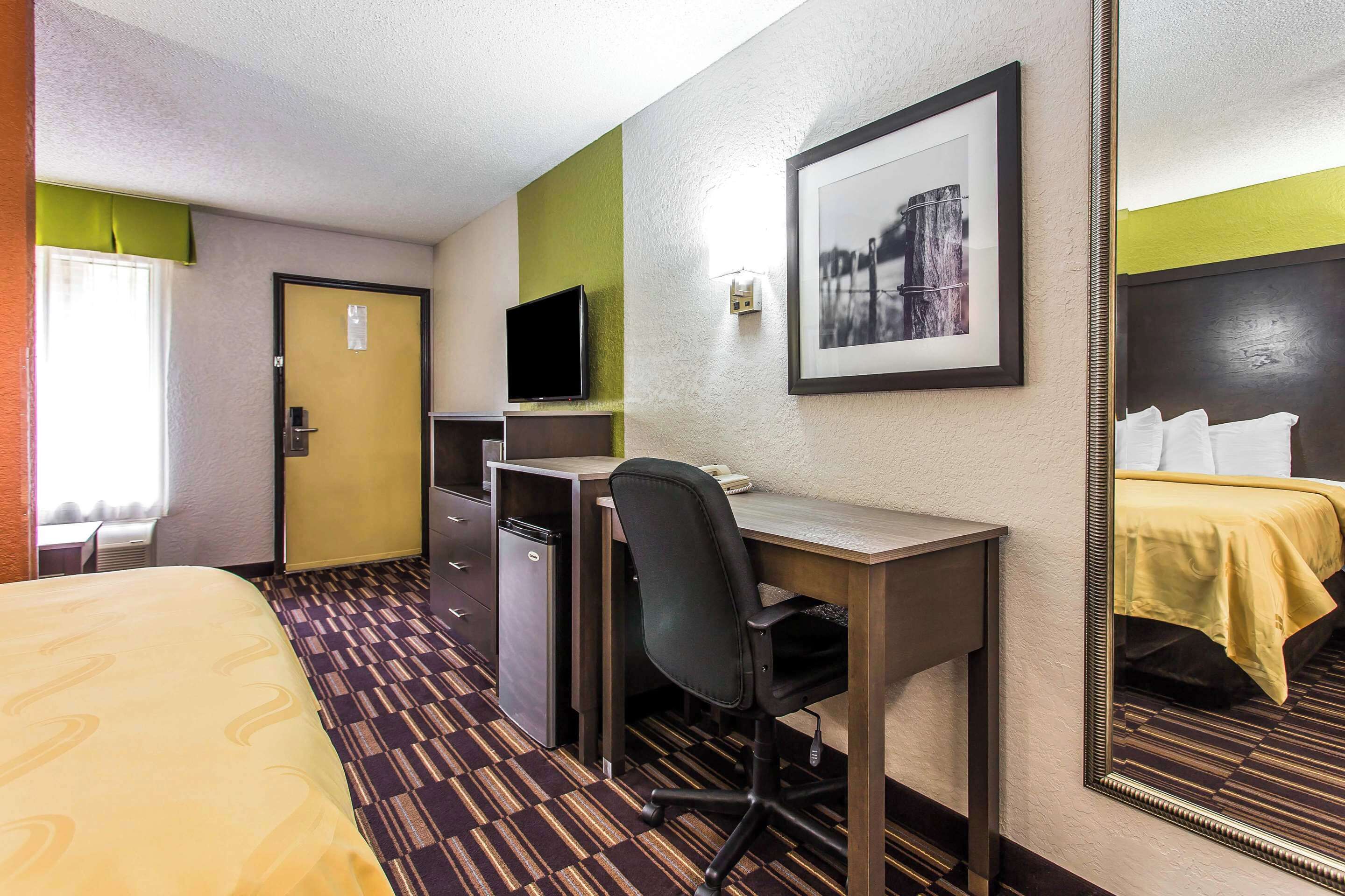 Habitación de hotel con cama, escritorio, silla, televisor y espejo.