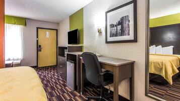Habitación de hotel con cama, escritorio, silla, televisor y espejo.