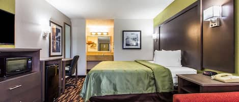 A hotel room with a bed, a desk with a telephone, a microwave, a refrigerator, a chair, and a red armchair.