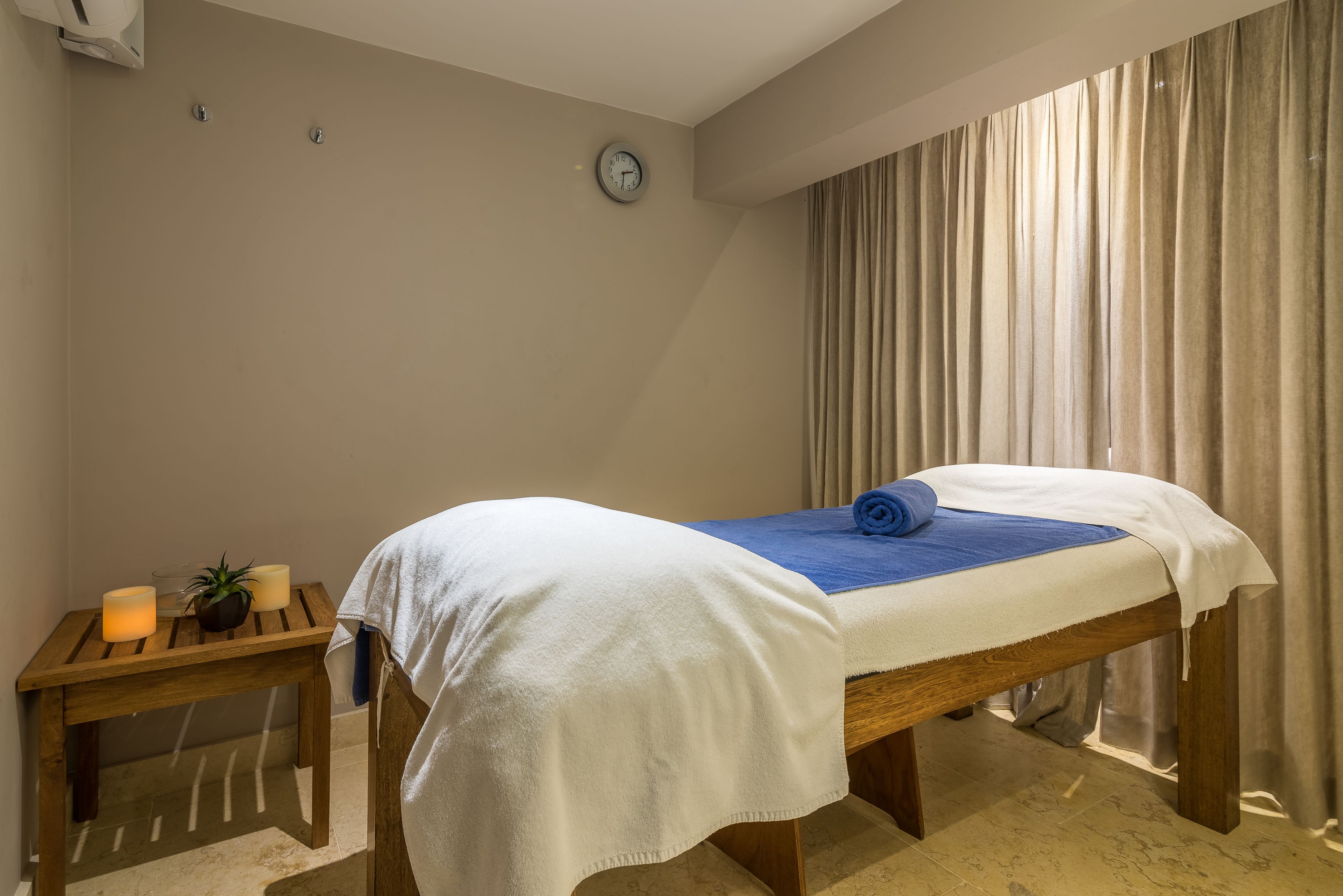 Un lit simple avec une couvre-lit blanche et bleue, une table de chevet en bois avec des bougies et une horloge au mur.
