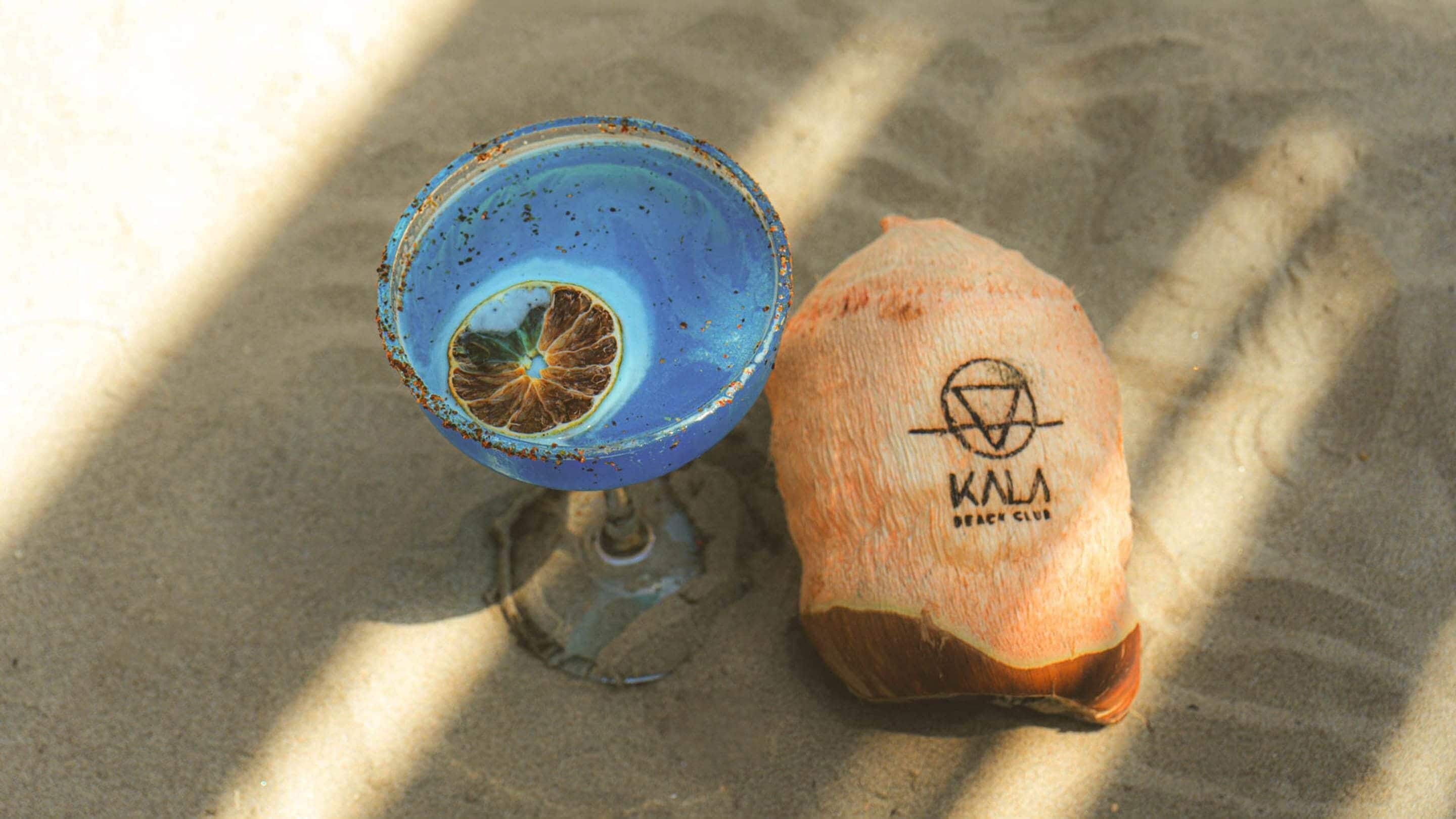 Ein blaues Glas mit einer getrockneten Orangenscheibe, eine Kokosnussmuschel mit einem Logo und Sand.
