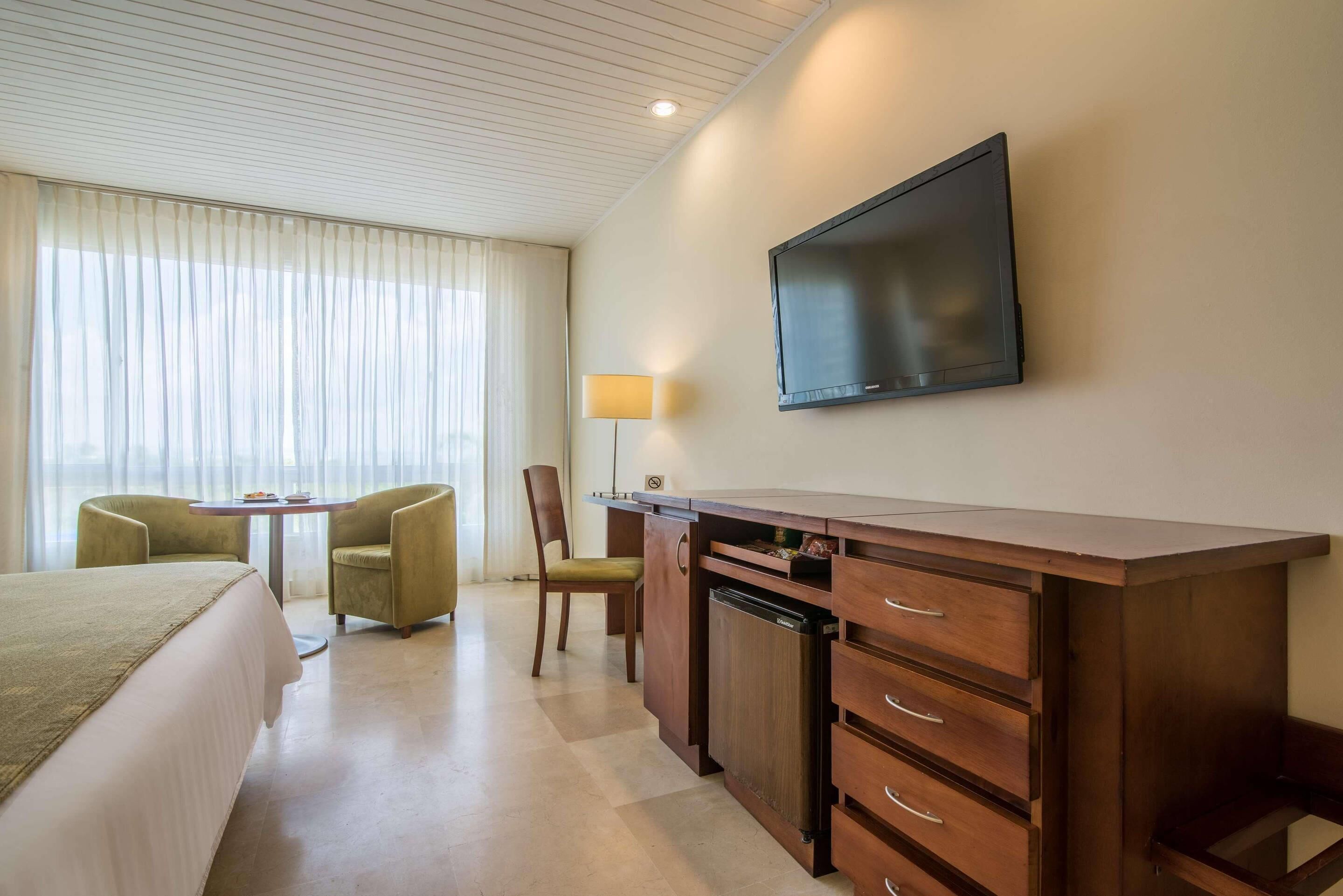 A hotel room with a bed, a desk with drawers, a television, a lamp, and a chair.