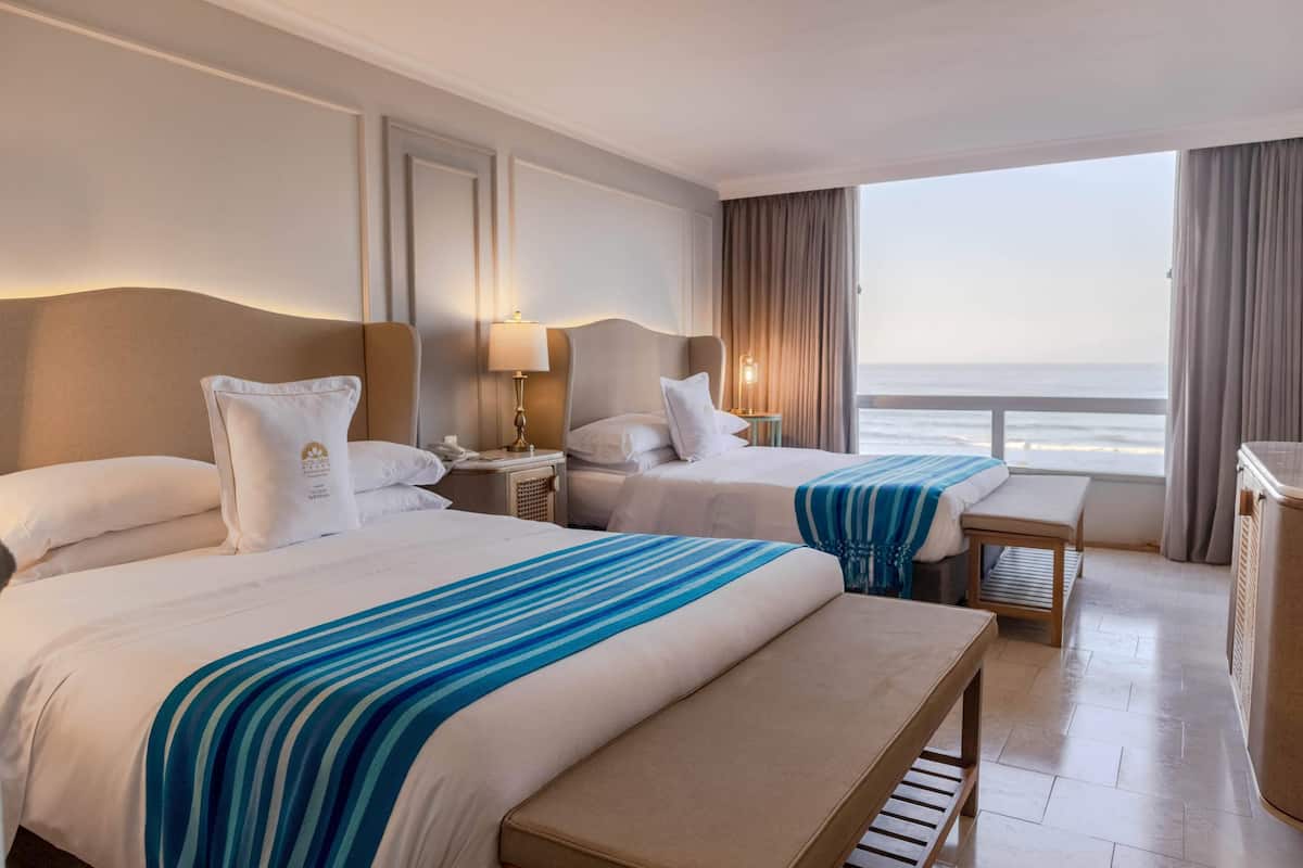 Een hotelkamer met twee bedden, een bankje, een nachtkastje, een lamp en uitzicht op de zee.