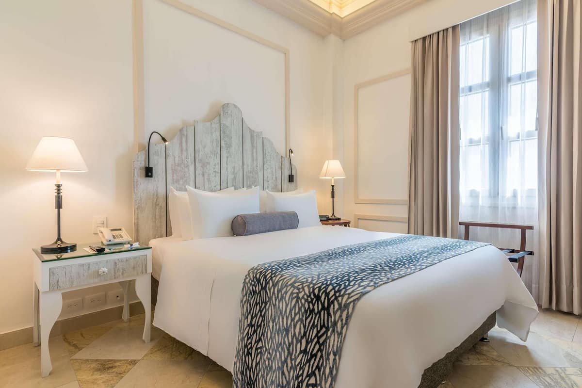 Een hotelkamer met een groot bed, twee nachtkastjes met lampen, een decoratief hoofdbord en uitzicht buiten door een raam met gordijnen.