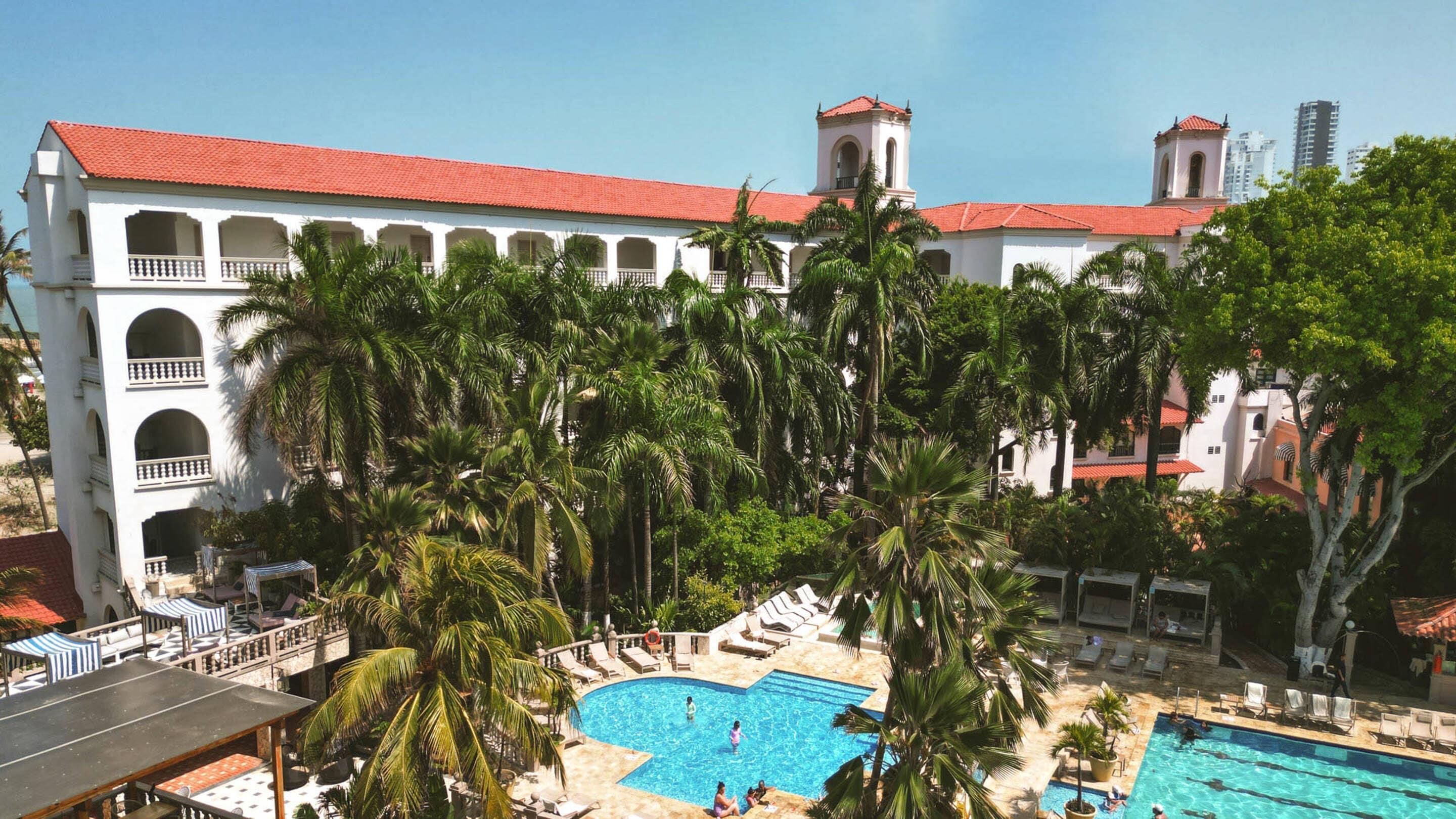 A hotel with a pool surrounded by palm trees, a red-roofed building, and a clear sky.