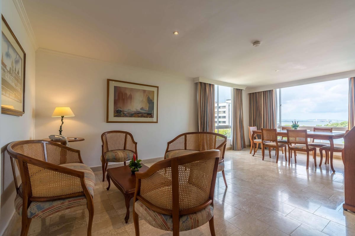 Een hotelkamer met een zithoek, rieten stoelen, een tafel, een lamp en een schilderij aan de muur.