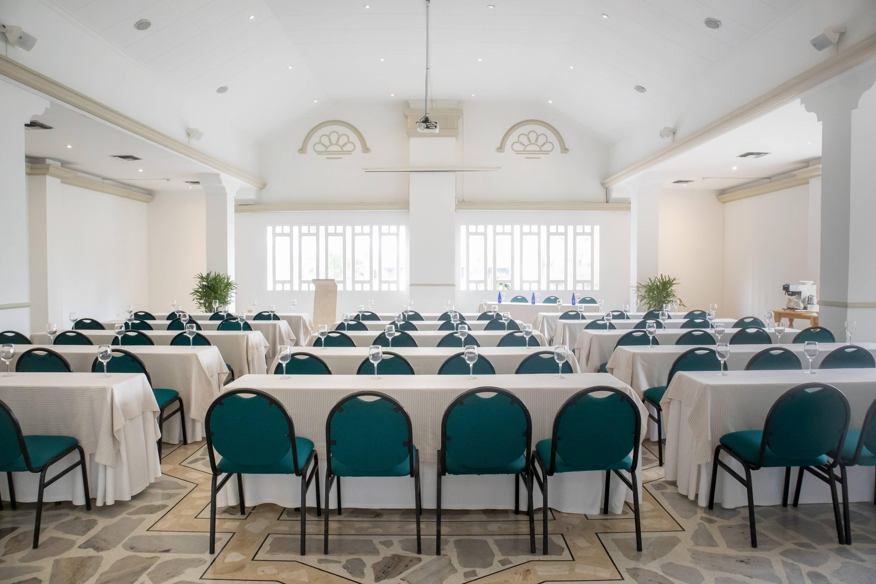 A large conference room with rows of tables and chairs, large windows, and a stone floor.