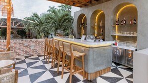 Zona de bar con suelo a cuadros, sillas de madera, mostrador de bar y estanterías con botellas y vasos.