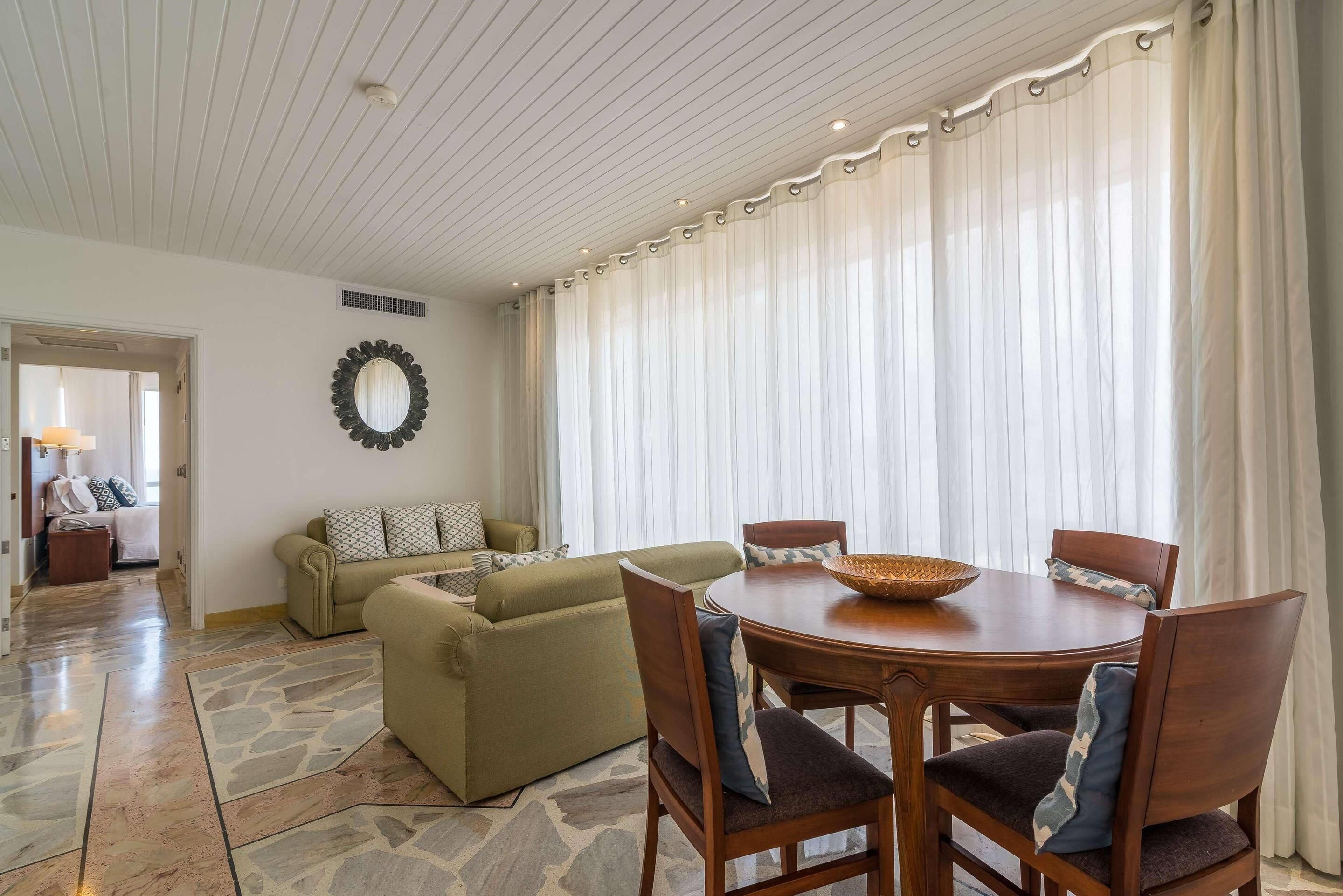 A living room with a dining area, stone flooring, a round wooden table, and a large window with curtains.
