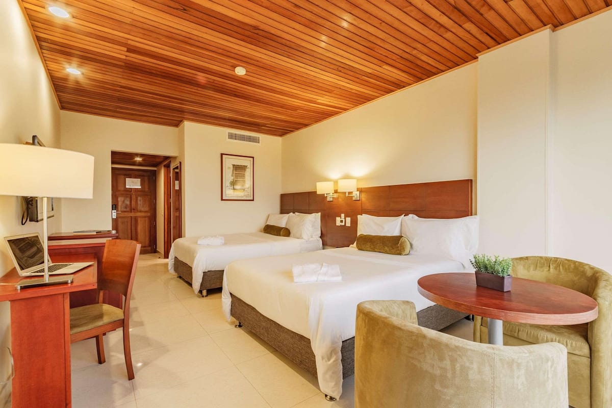 Een hotelkamer met twee bedden, een bureau met een laptop, een stoel, een ronde tafel met een plant en een houten plafond.