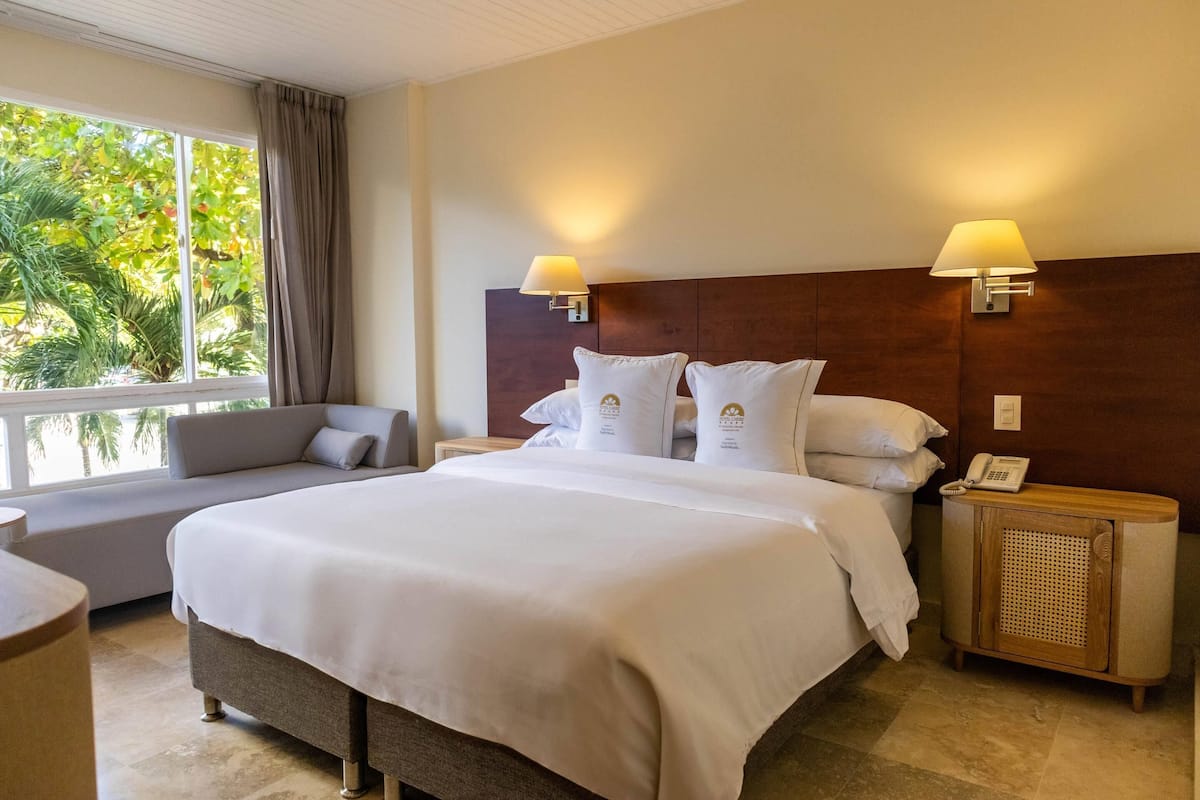 Een hotelkamer met een groot bed, twee bedlampen, een nachtkastje met een telefoon en een raam met uitzicht op groen.