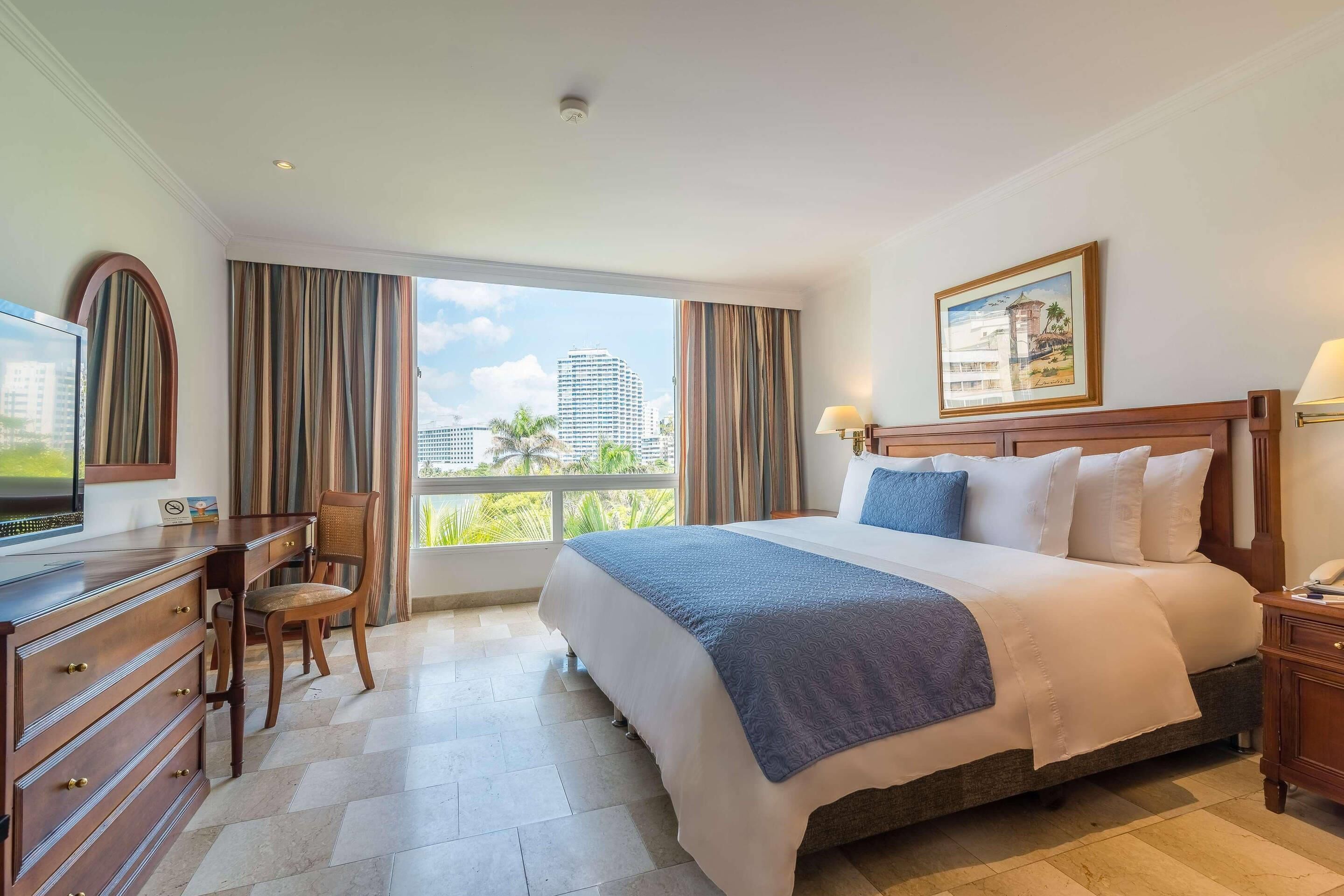 A hotel room with a large bed, a desk with a chair, a television, and a window with curtains.