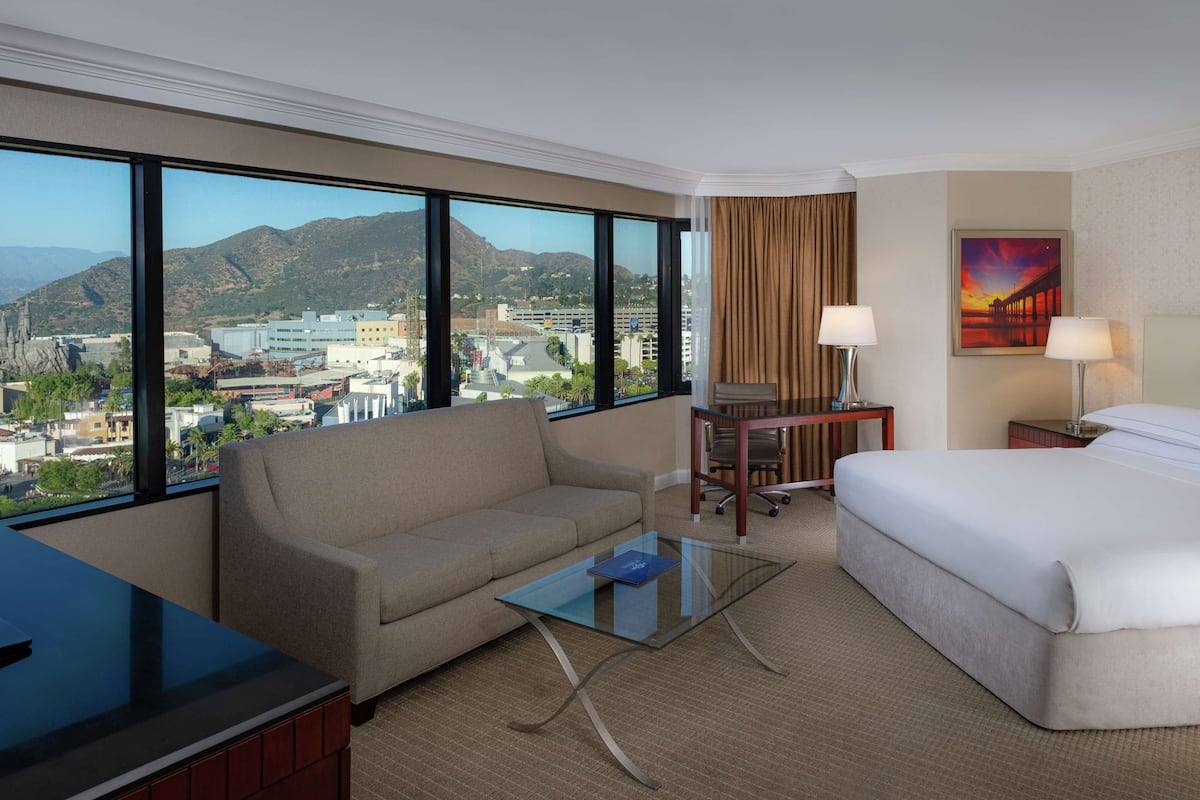 A hotel room with a large window, a sofa, a bed, a glass coffee table, and a desk with a lamp.