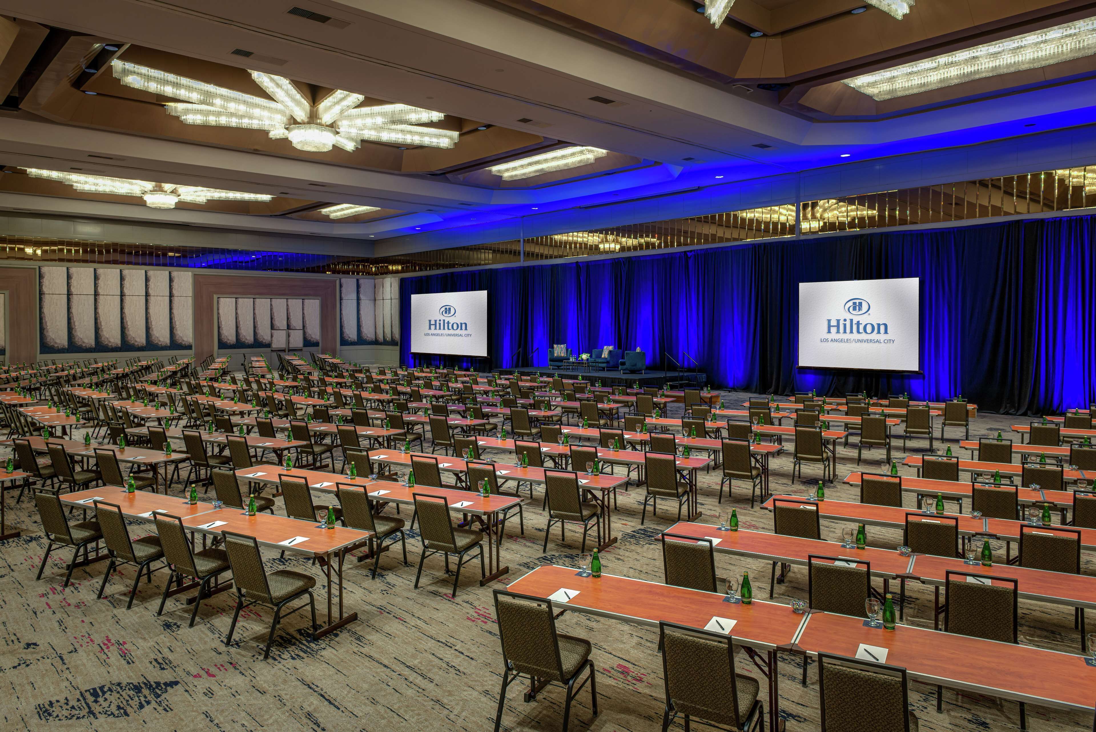 A large conference room with rows of tables and chairs, a stage with a blue curtain backdrop, and two large screens displaying the Hilton logo.