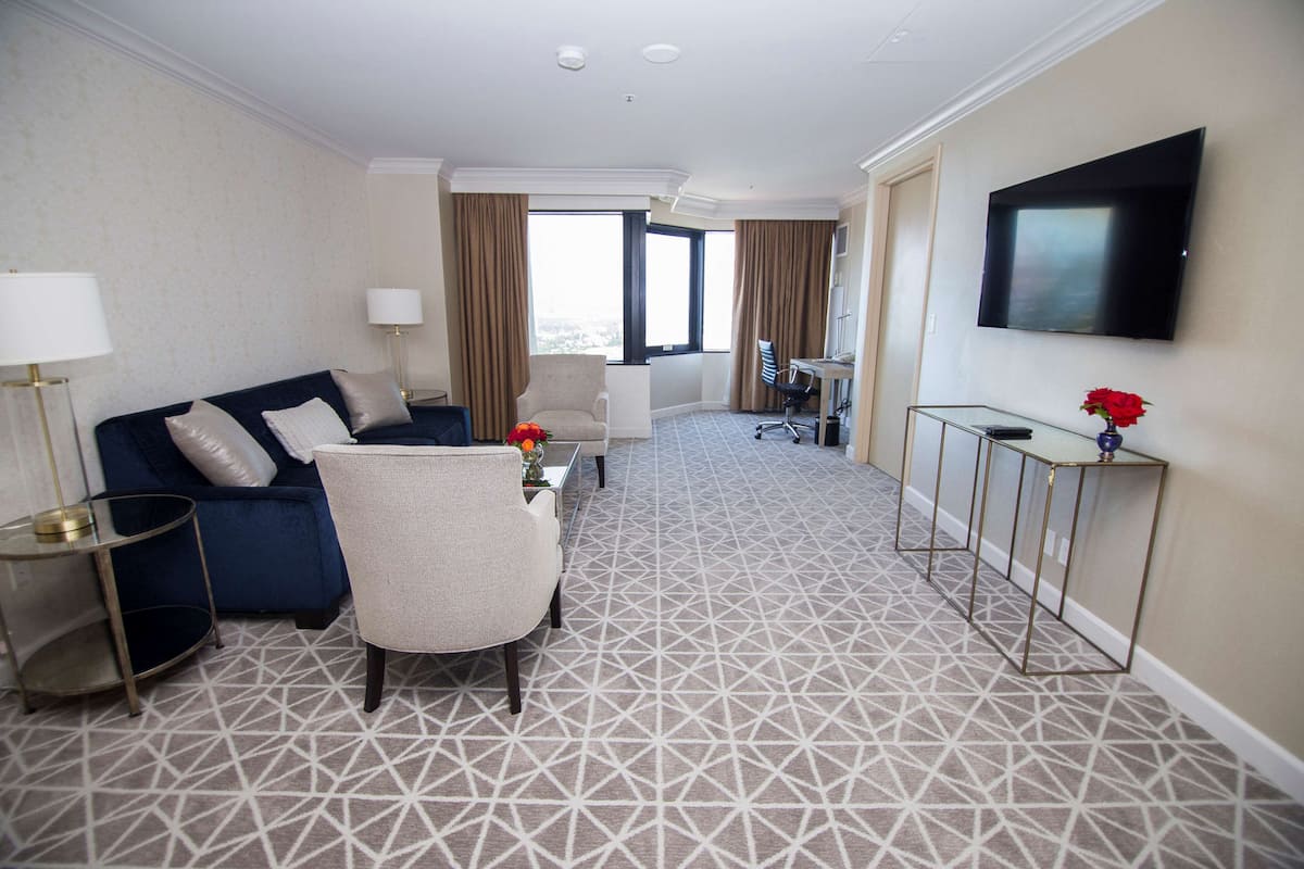 A hotel room with a blue sofa, a beige armchair, a glass coffee table, a TV mounted on the wall, and a patterned carpet.