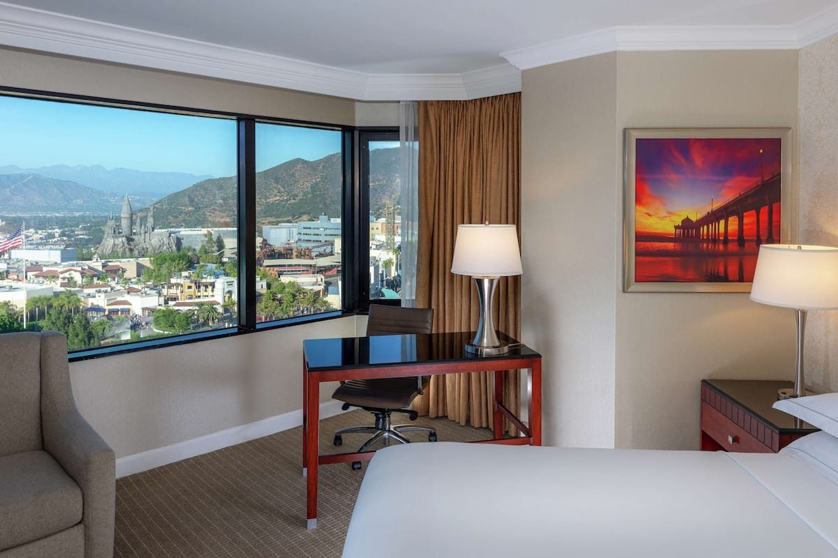 A hotel room with a large window offering a city view, a desk with a chair, a bed, and a painting on the wall.