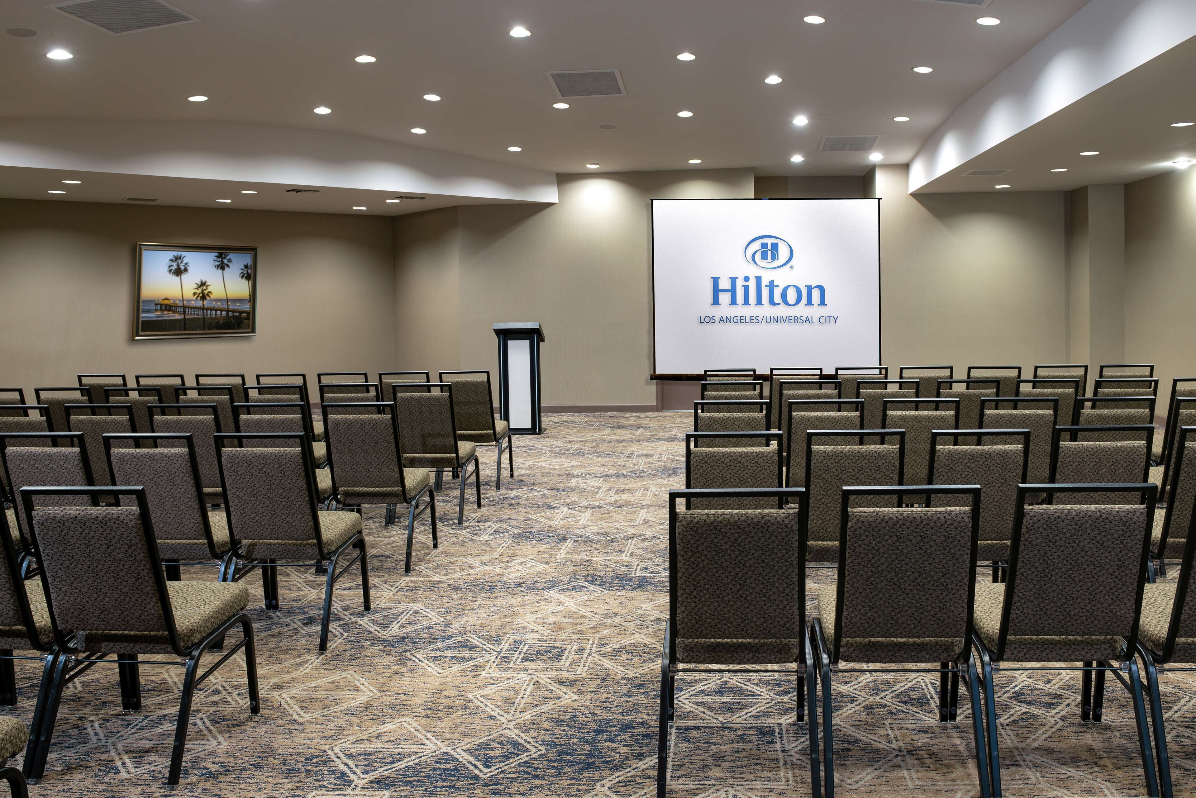 A conference room with rows of chairs, a podium, and a large screen displaying the Hilton logo.
