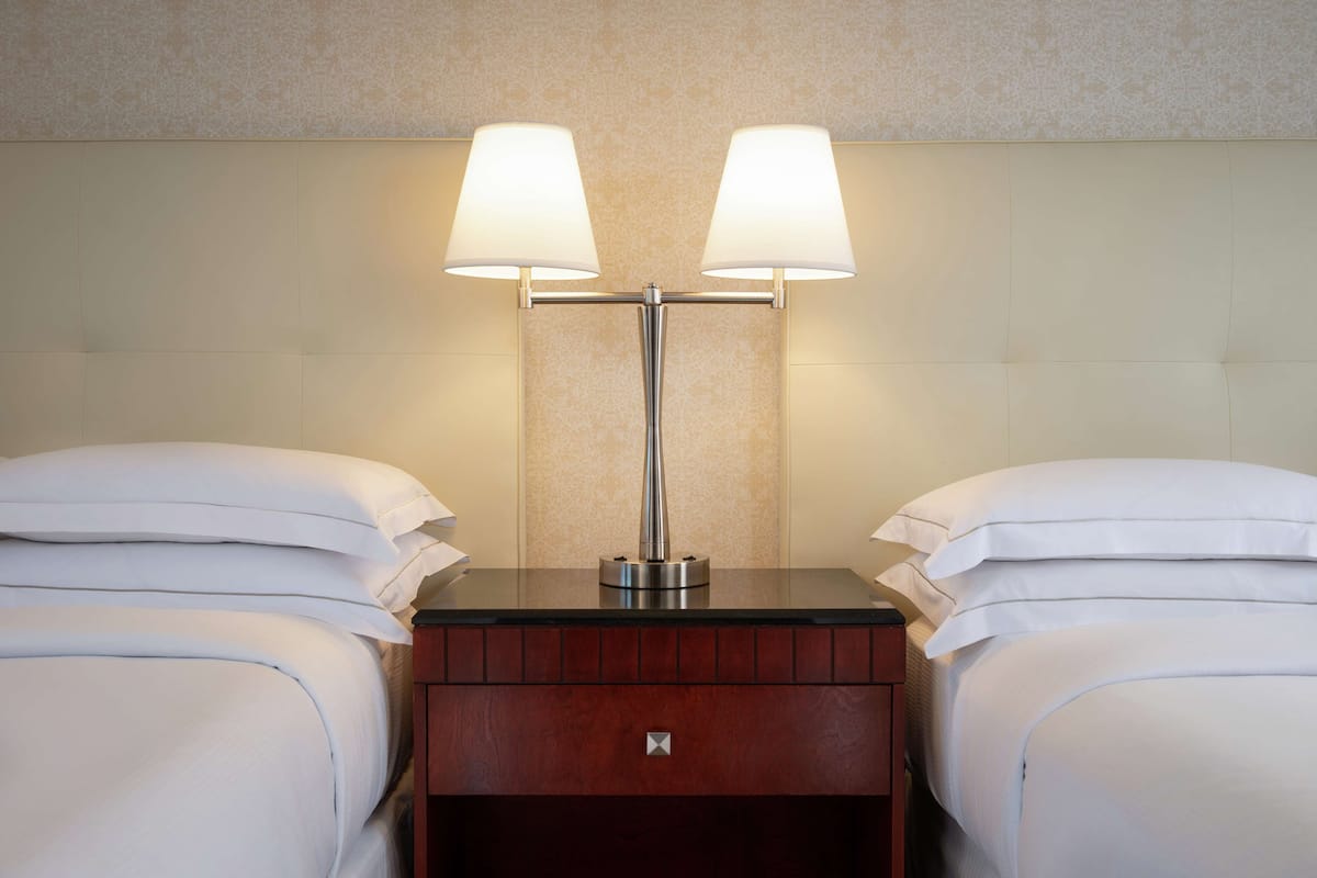 A hotel room with two beds, a nightstand, and a lamp between them.
