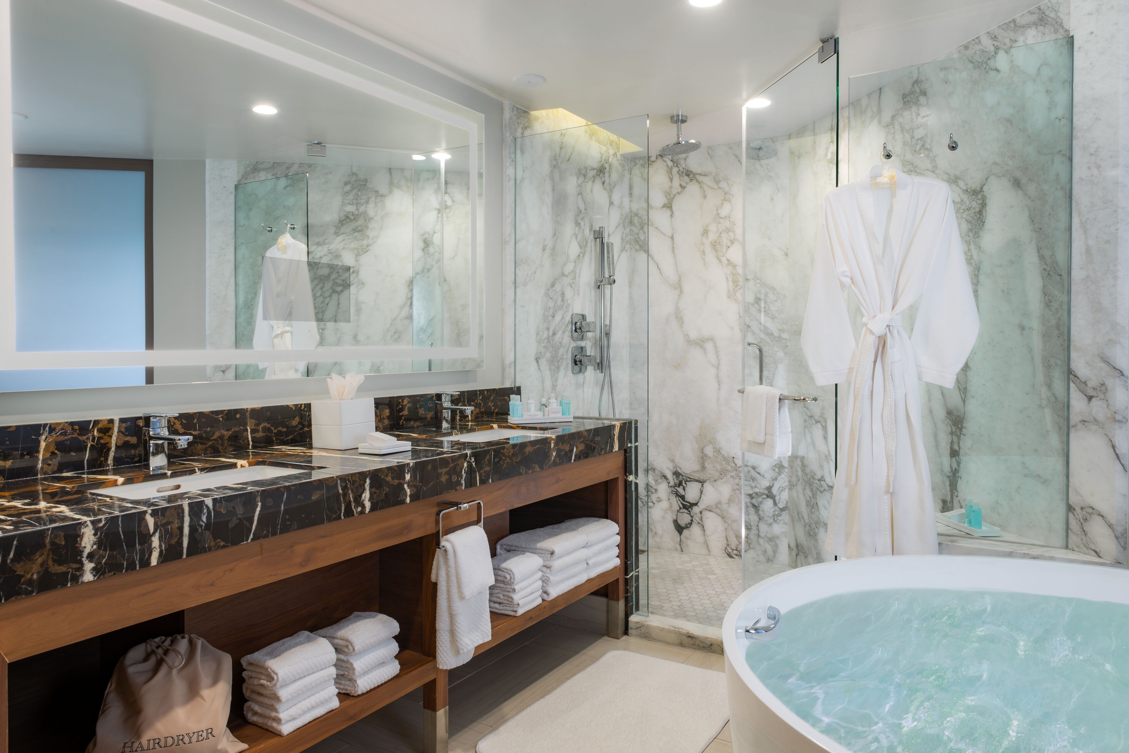 A modern bathroom with a marble wall, a large mirror, a vanity with a black marble countertop, a bathtub, and a shower area.