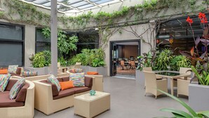 Un área de descanso interior y exterior con mobiliario de mimbre, plantas en macetas y techo de vidrio.