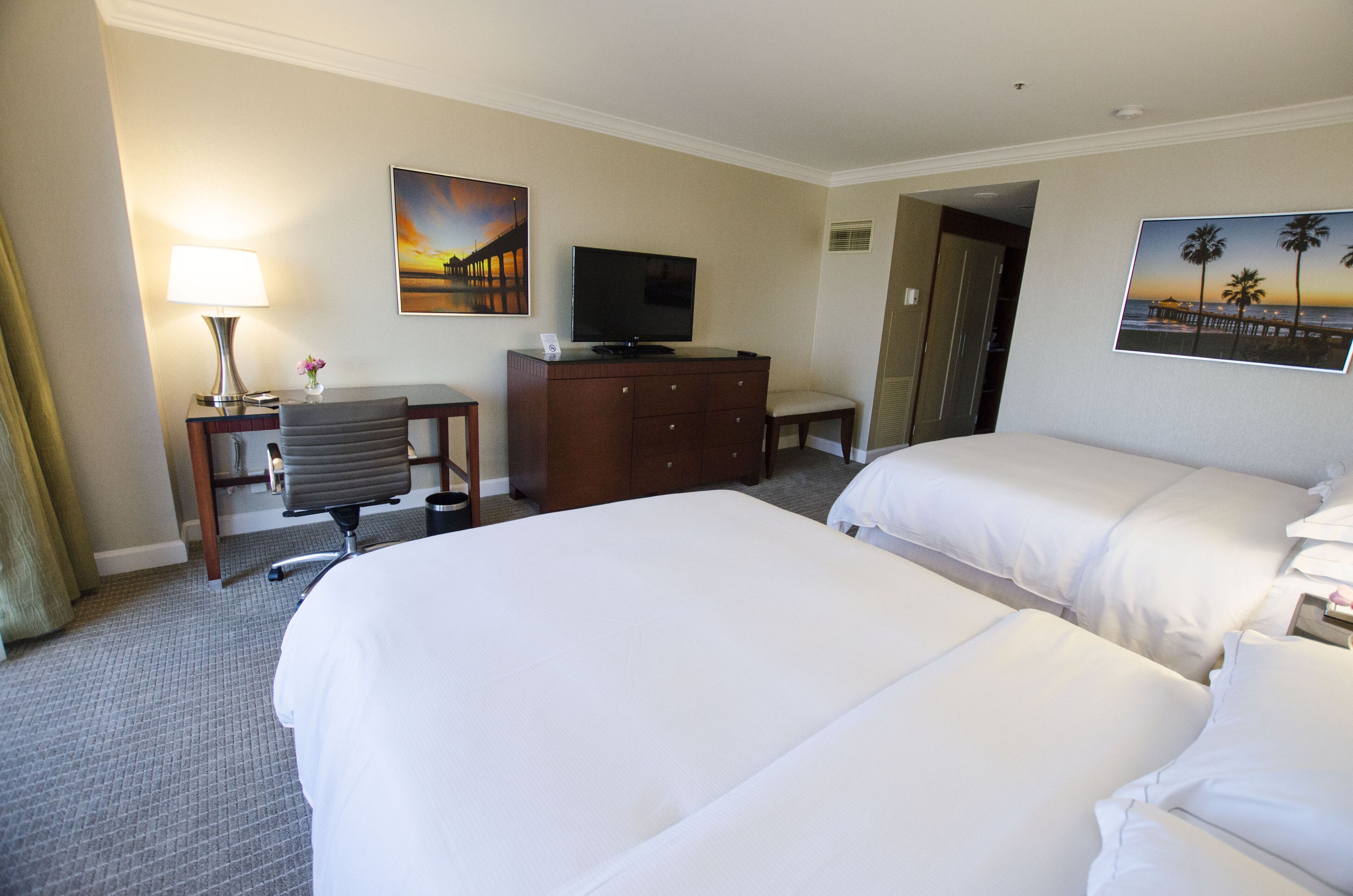 Quarto de hotel com duas camas, uma escrivaninha com abajur, televisão e obras de arte nas paredes.