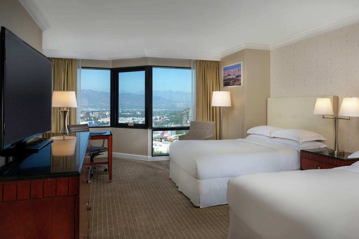 A hotel room with a large TV, two beds, a desk with a chair, and a view of a cityscape through large windows.