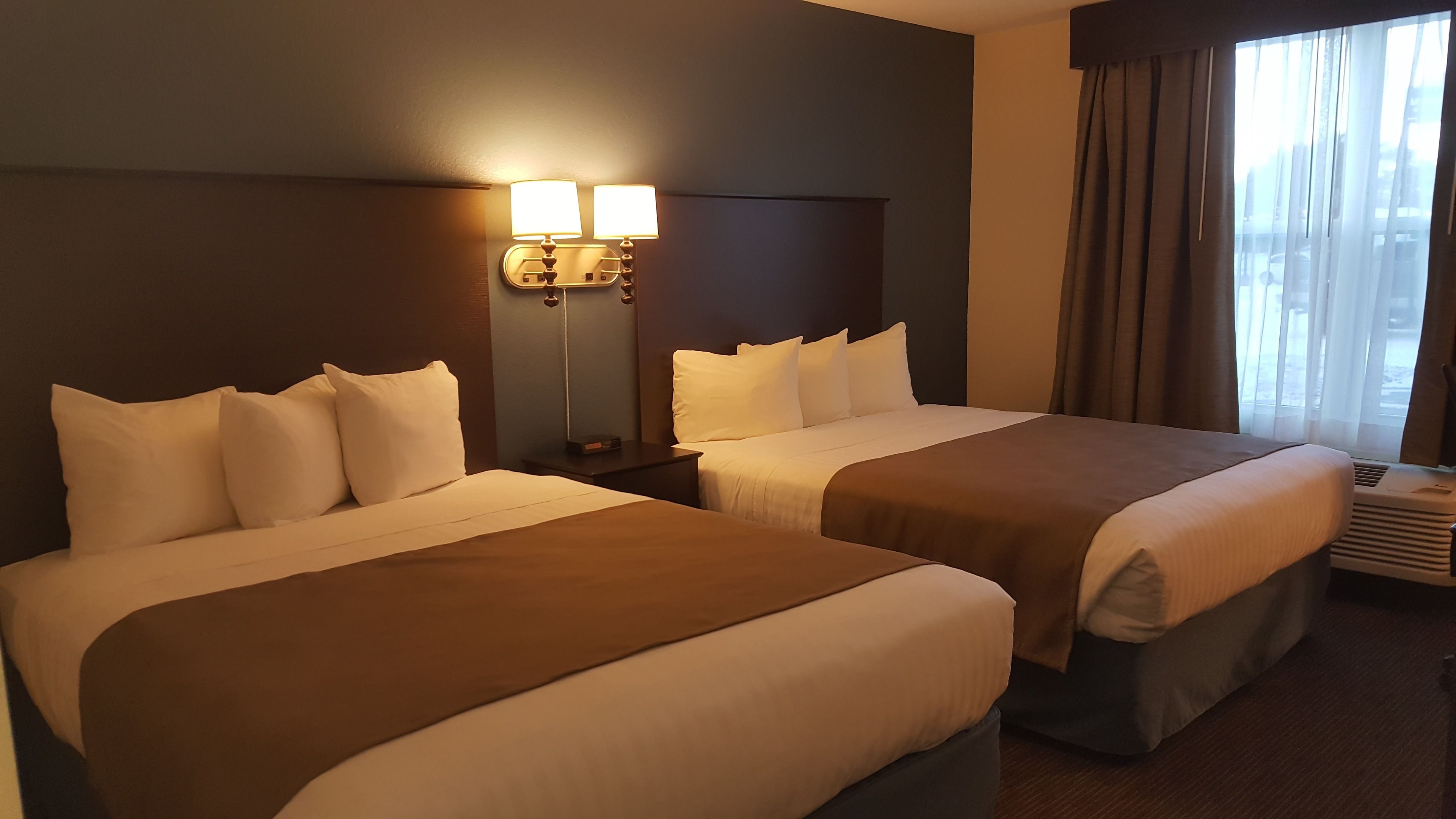 A hotel room with two beds, a nightstand, a lamp, and a window with curtains.