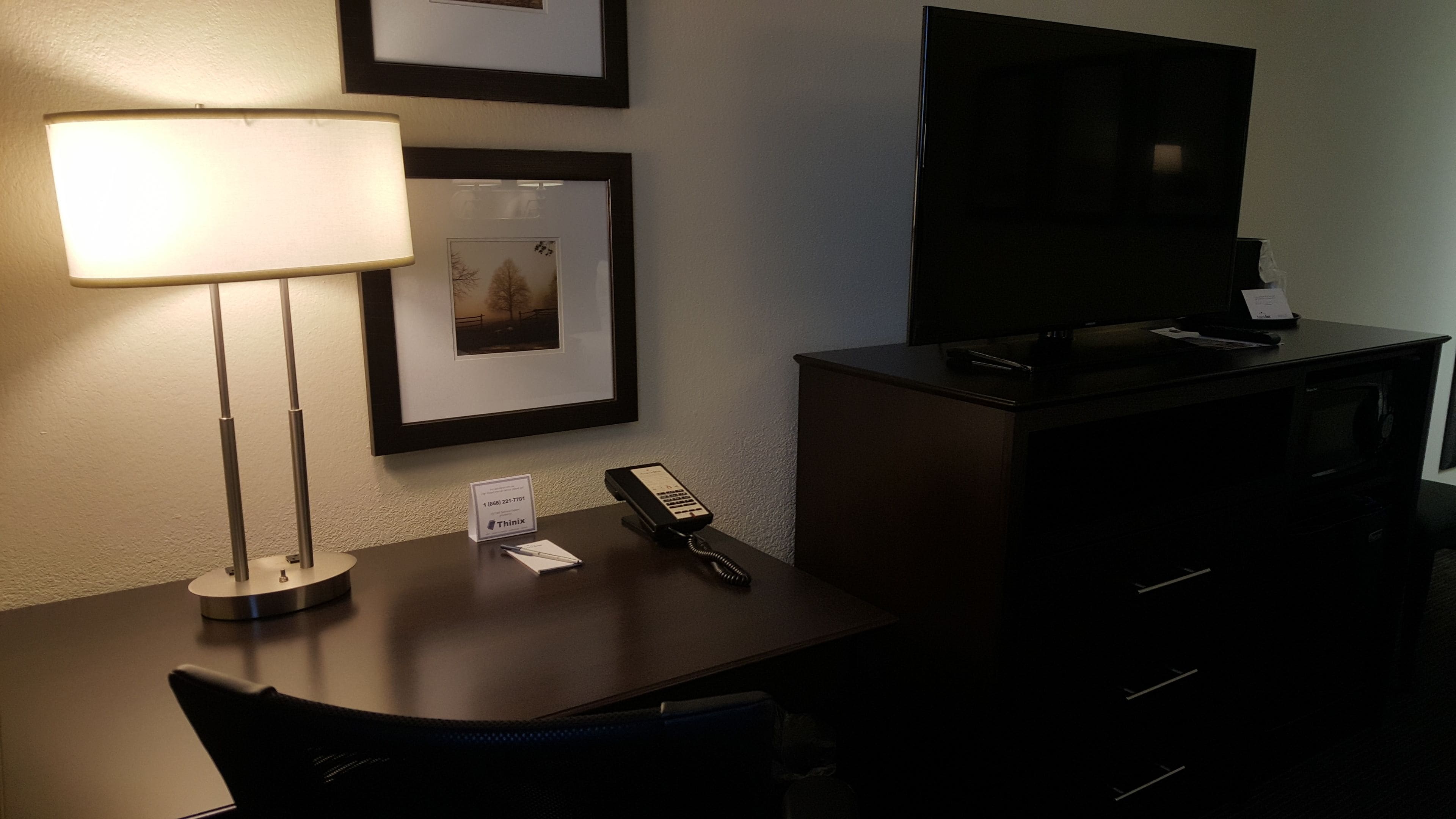 A hotel room with a desk, a lamp, a telephone, a television, and a framed picture on the wall.