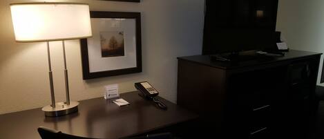 A hotel room with a desk, a lamp, a telephone, a television, and a framed picture on the wall.
