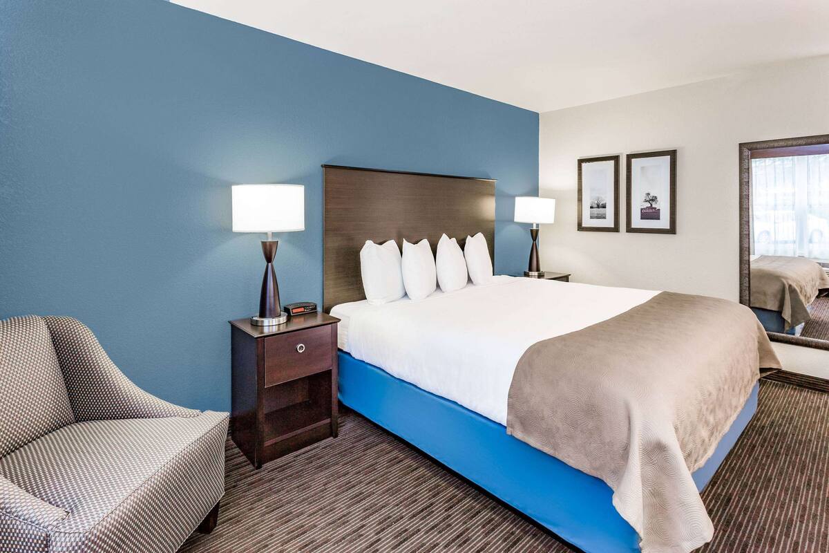 Una habitación de hotel con una cama grande, una mesita de noche con lámpara, una silla y un espejo.