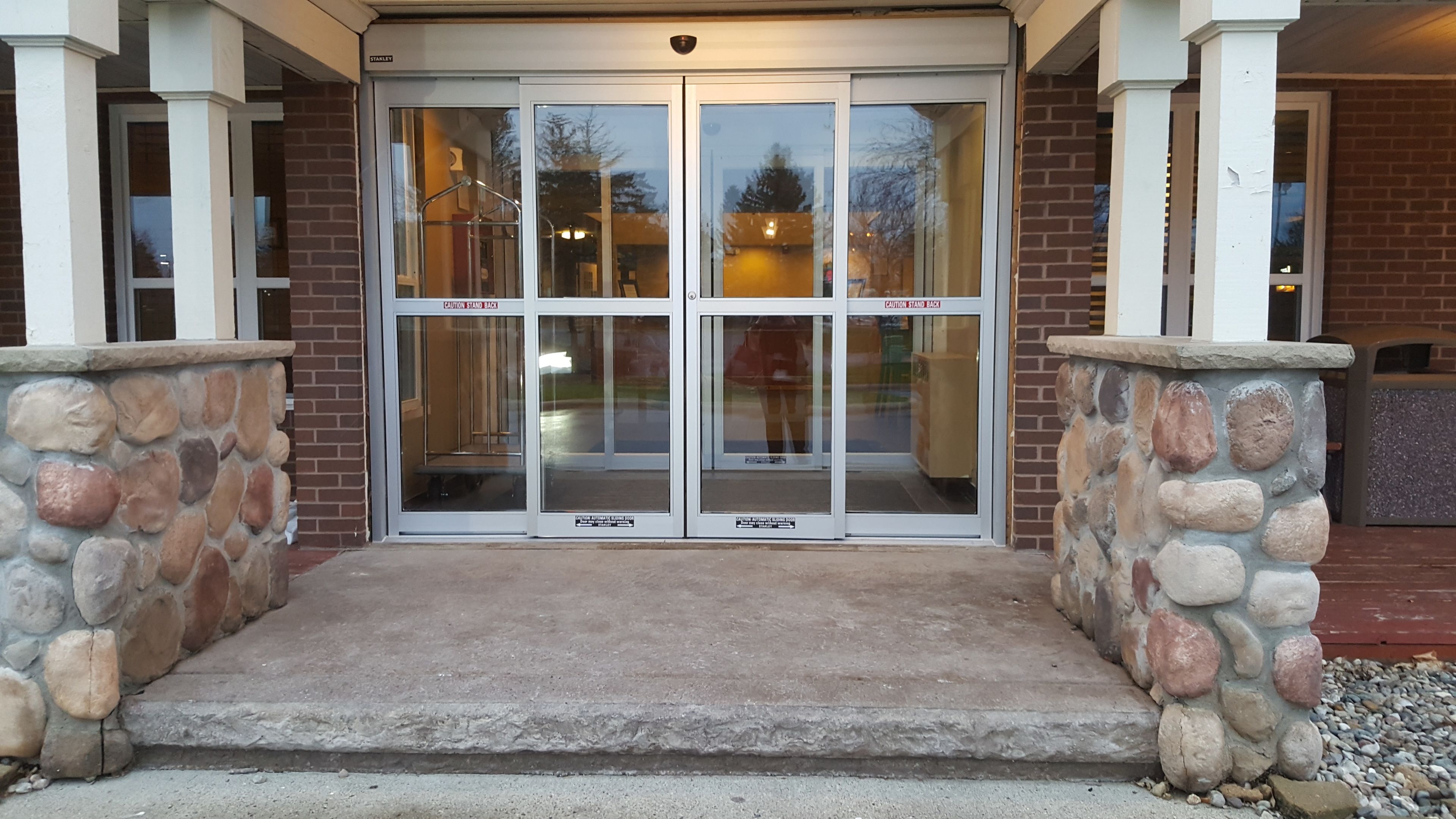 A building entrance with a stone wall, a concrete step, and a glass door with a "Caution Stand Back" sign.
