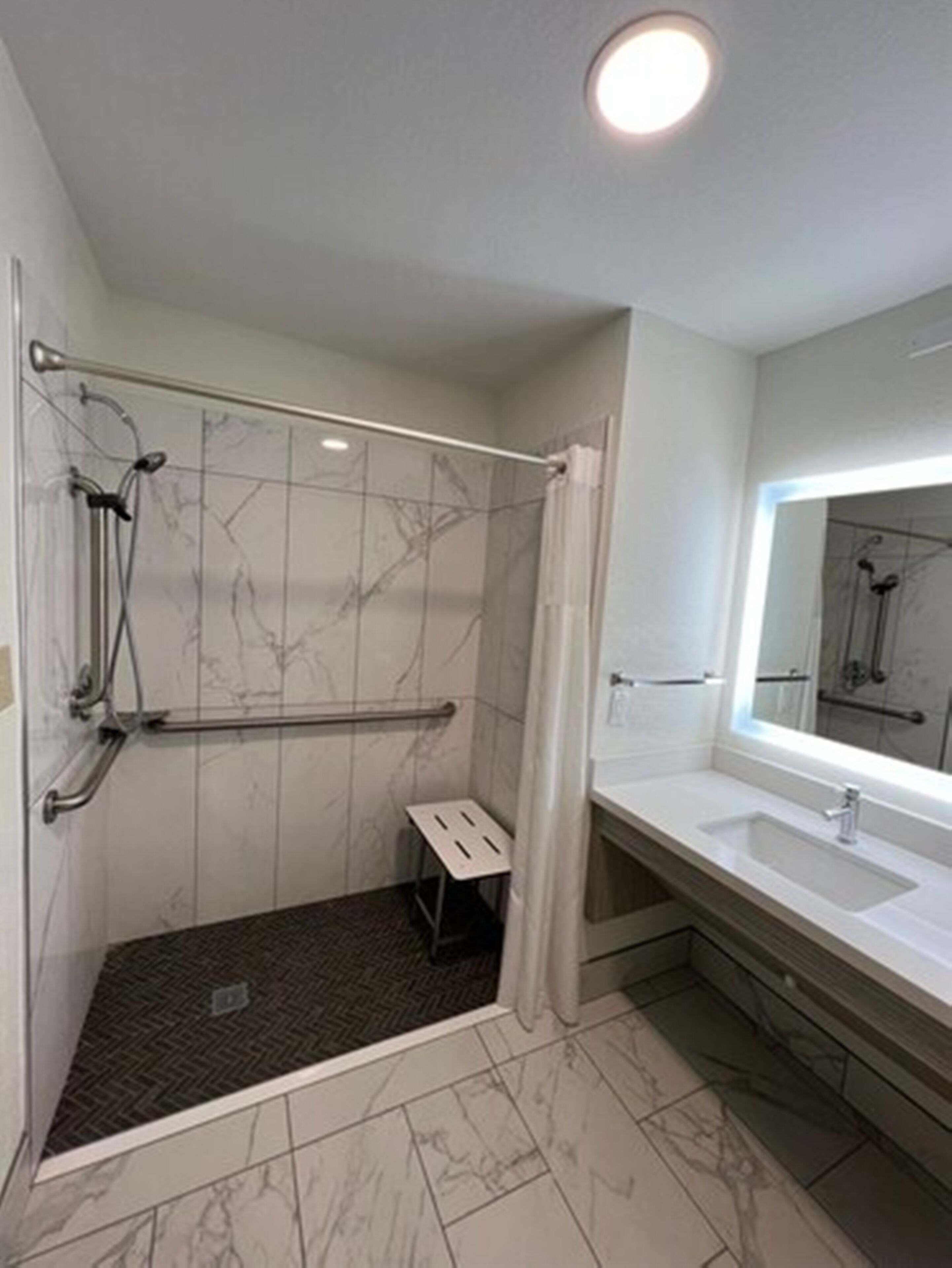 Un baño moderno con ducha a nivel, un taburete pequeño, un espejo grande y un mueble de tocador con lavamanos.