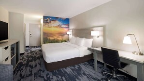 A hotel room with a bed, desk, chair, TV, and a painting of a sunset.