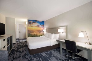 A hotel room with a bed, desk, chair, TV, and a painting of a sunset.