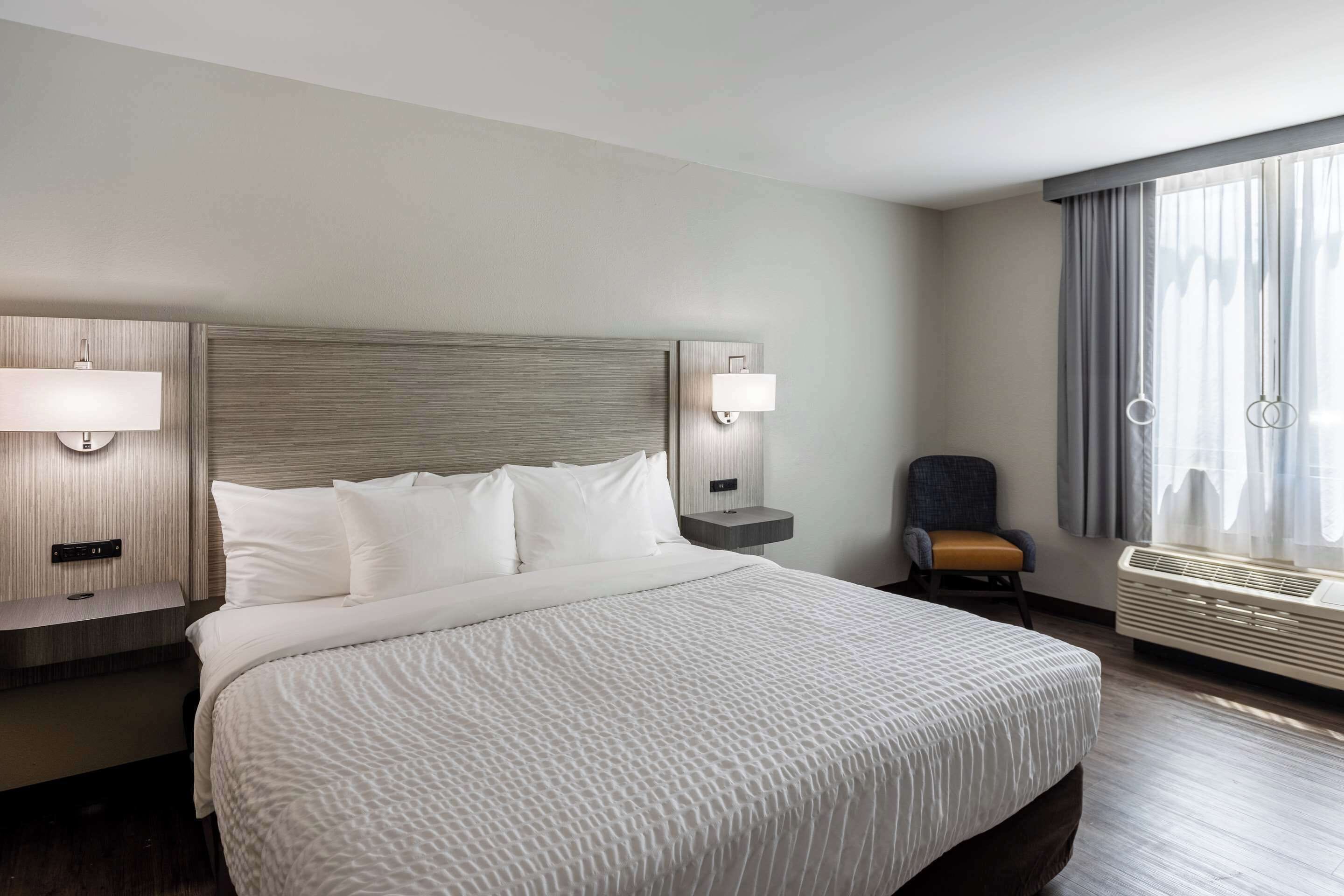 Ein Hotelzimmer mit einem großen Bett, zwei Nachttischen mit Lampen, einem Sessel und einem Fenster mit Vorhängen.
