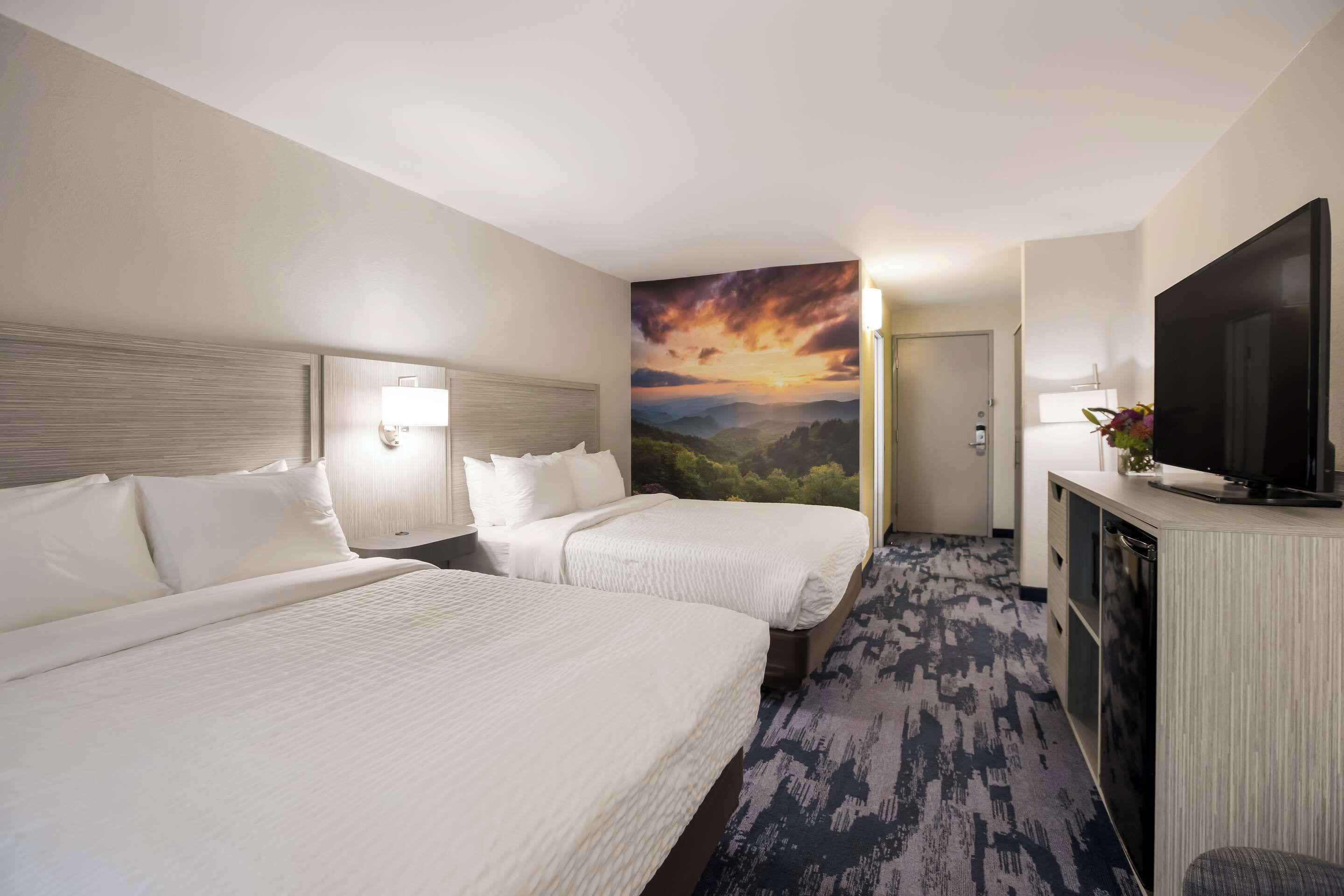 A hotel room with two beds, a TV, a desk, and a painting of a sunset.