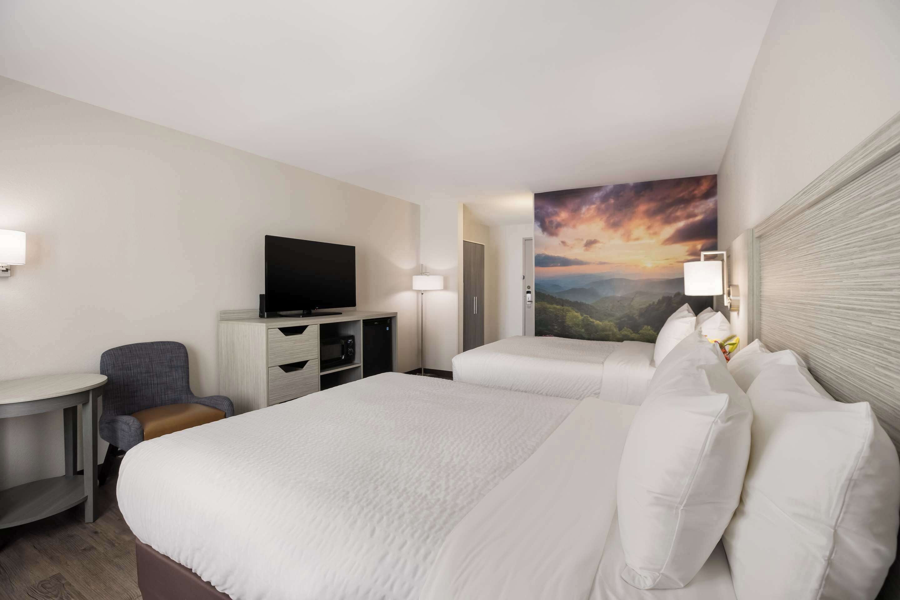 A hotel room with a large bed, a desk with a chair, a television, and a painting of a sunset on the wall.