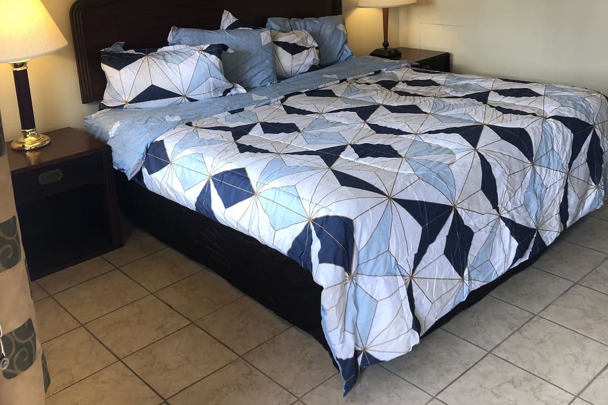 A neatly made bed with a geometric patterned duvet, two bedside lamps, and a framed painting above the bed.
